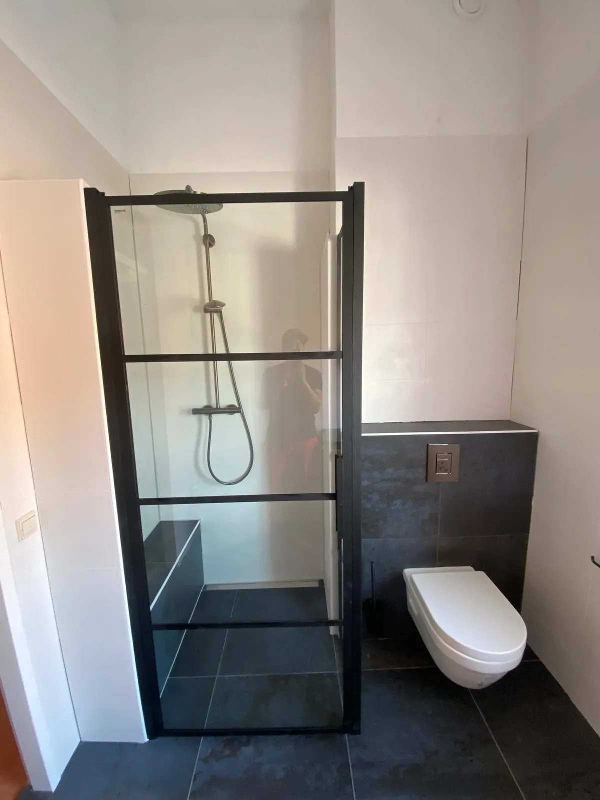 Inloopdouche met zwarte glazen deur, regendouche, getegelde zitbank en hangend toilet in een voorzetwand